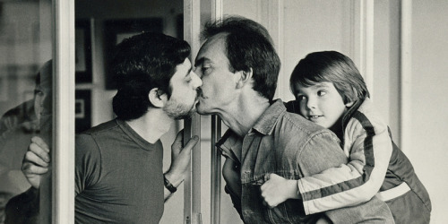 a silver-tinted photograph of a man leaning out a sliding door to kiss his husband, who has their young daughter clinging to his back