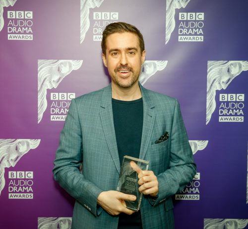 A photo of a smiling and suited Robert Valentine, posing with a BBC Audio Drama Award in front of a BBC Audio Drama Awards backdrop