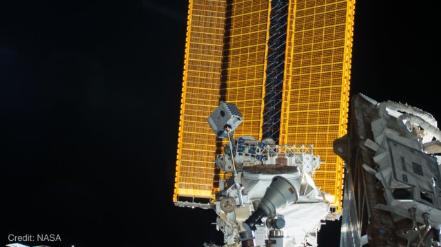 This photo shows the NICER payload on the International Space Station. Against a black background, tall rectangular solar panels that appear as a golden mesh rise from the bottom of the photo, passing through its middle area. In front of that are a variety of gray and white shapes that make up instruments and the structure of the space station near NICER. Standing above from them, attached to a silver pole, is the rectangular box of the NICER telescope, which is pointing its concentrators up and to the right. Credit: NASA.