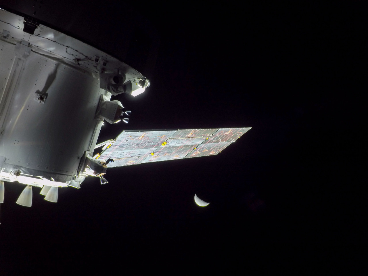 A cross-section of the white, cylindrical Orion spacecraft dominates the left side of the screen. A rectangular solar array snaked with multicolored wires extends up and out to 2 o'clock, and four bell-shaped auxiliary thrusters point down from the body of the spacecraft. The small crescent Moon is isolated in the black of space in the distance beyond and below the solar array. Credit: NASA