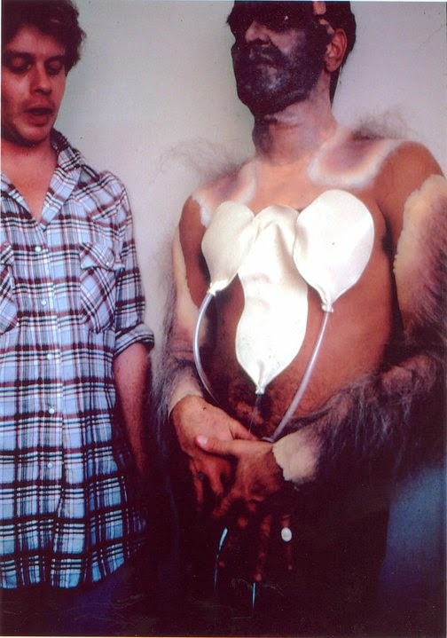 Two men standing side by side, one wearing werewolf prosthetics and inflatable latex balloons on his chest