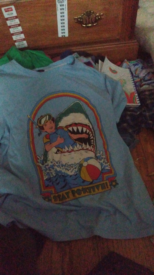 A blue tee with a graphic of a kid being eaten by a shark while giving a thumbs up and text saying "Stay positive"