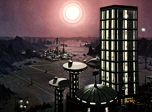 GIF 6: scenic view of a more rural landscape at twilight. in the foreground is a tall high rise building with large windows, two towers, and a smaller building with a dome roof. all the buildings are illuminated from inside. further in the background is another group of buildings with a crane in its center. there are mountain ranges on either side of the frame while the center with the two city areas is flat land. the sky is pink and a sun is hanging in the sky, either half risen or half set (I don't know lol).
