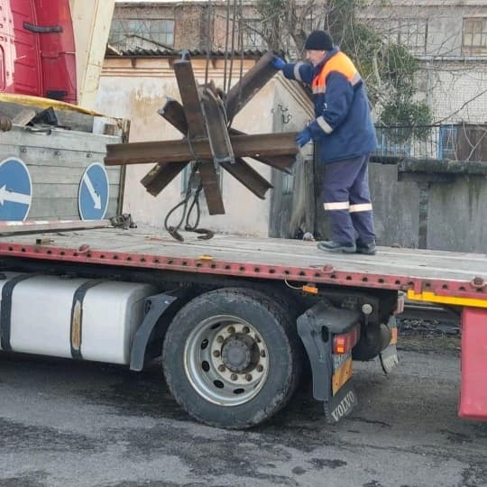 A man in high vis stands on the back of a flat bed truck, wrapping chains around a piece of anti-tank equipment. It seems to be multiple iron girders welded together into a many-limbed star, and is almost as tall as the man.