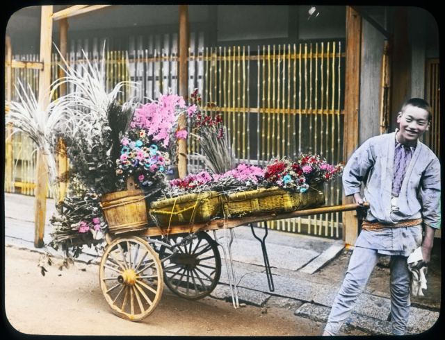 A young boy is standing by a cart full of flowers in baskets. He is wearing a traditional workmen’s clothing and smiling. Flowers are painted vividly.