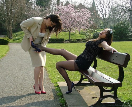 Two ladies wearing high heels and pantyhose teases on a bench in a park