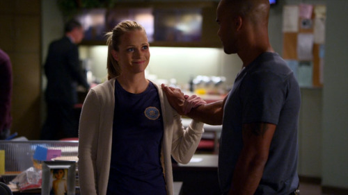 In an office, Derek Morgan, a man with a shaved head wearing a blue-grey shirt, has his hand on the shoulder of JJ, a blonde woman wearing a white sweater over a blue FBI shirt. She is smiling and holding his wrist. She has a black eye.