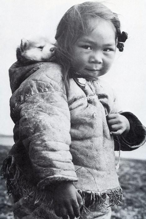 darksilenceinsuburbia:An Inuk girl with her husky puppy in the hood of her amatiuq circa 1920. Mothers would also carry their infants in the same manner. This little girl is playing mommy and baby with her puppy.