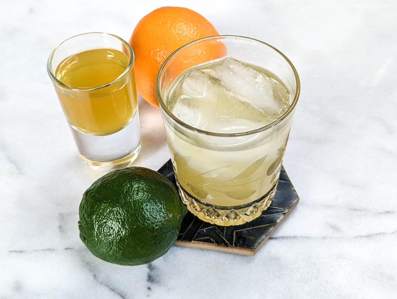 Three-quarter photograph of a margarita in a crystal glass on a hexagonal coaster, with a lime next to it and a shot glass of orange liqueur next to an orange behind them, all on a marble background