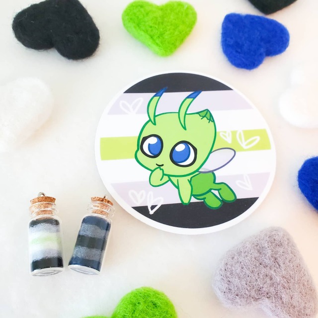 a photo of various agender and gendervoid items including needle felted hearts, two bottle charms, and a sticker. The sticker has the pokemon celebi and the agender flag on it.