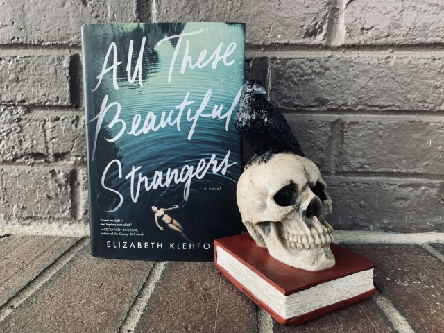 Shown is ALL THESE BEAUTIFUL STRANGERS (with cover art depicting a young woman swimming) against a backdrop of bricks, with a bookend in the form of a skull and raven sitting on top of an old book. Photo by AHS.