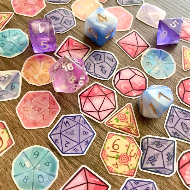 Product photo of individual flakes of pastel-colored dice stickers. These stickers include dice themed after candy, cherry blossoms, lavender, and roses. Actual pink, purple, and blue dice are scattered amongst the stickers to show how close in size the stickers are to real dice. 