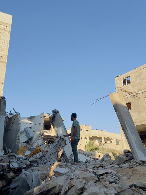 A photo of Hossam. He is dressed in the same clothes as the previous photo. The photo is taken from farther away. Hossam is facing away from the camera and standing on top of the rubble of a building.