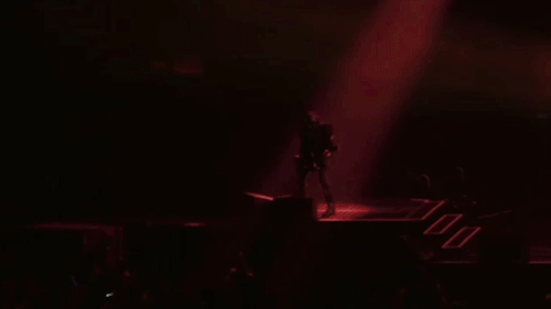 A distant shot of Phantom playing guitar on stage. A red spotlight illuminates him as he puts a foot on the monitor and lifts his guitar forward while playing.
