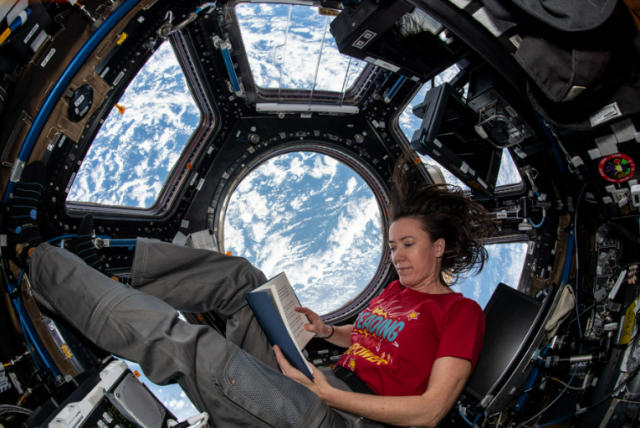 NASA astronaut Megan McArthur reads a blue book while floating in the cupola observation module on the International Space Station. She is wearing a red shirt and gray pants. Behind her, Earth can be seen through the module windows. Credit: NASA/Megan McArthur