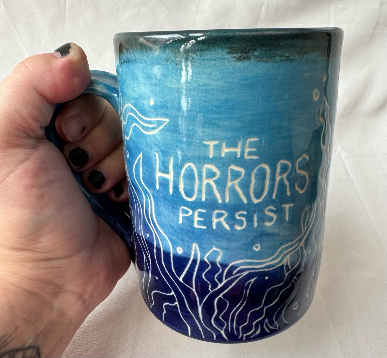 a turquoise and dark blue mug that reads 'the horrors persist' surrounded by carvings of kelp