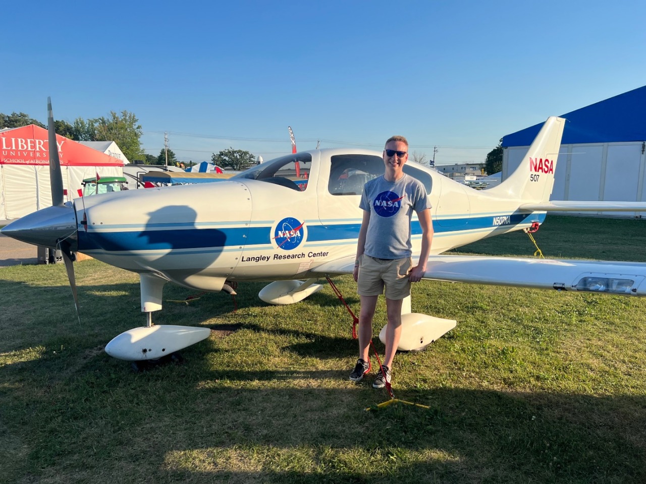 Adult Michael posing in front of a NASA aircraft at EAA Airventure 2022 in Oshkosh, WI. An adult man wearing a NASA t-shirt stands in front of a white and blue emblazoned propeller plane on a green patch of grass.