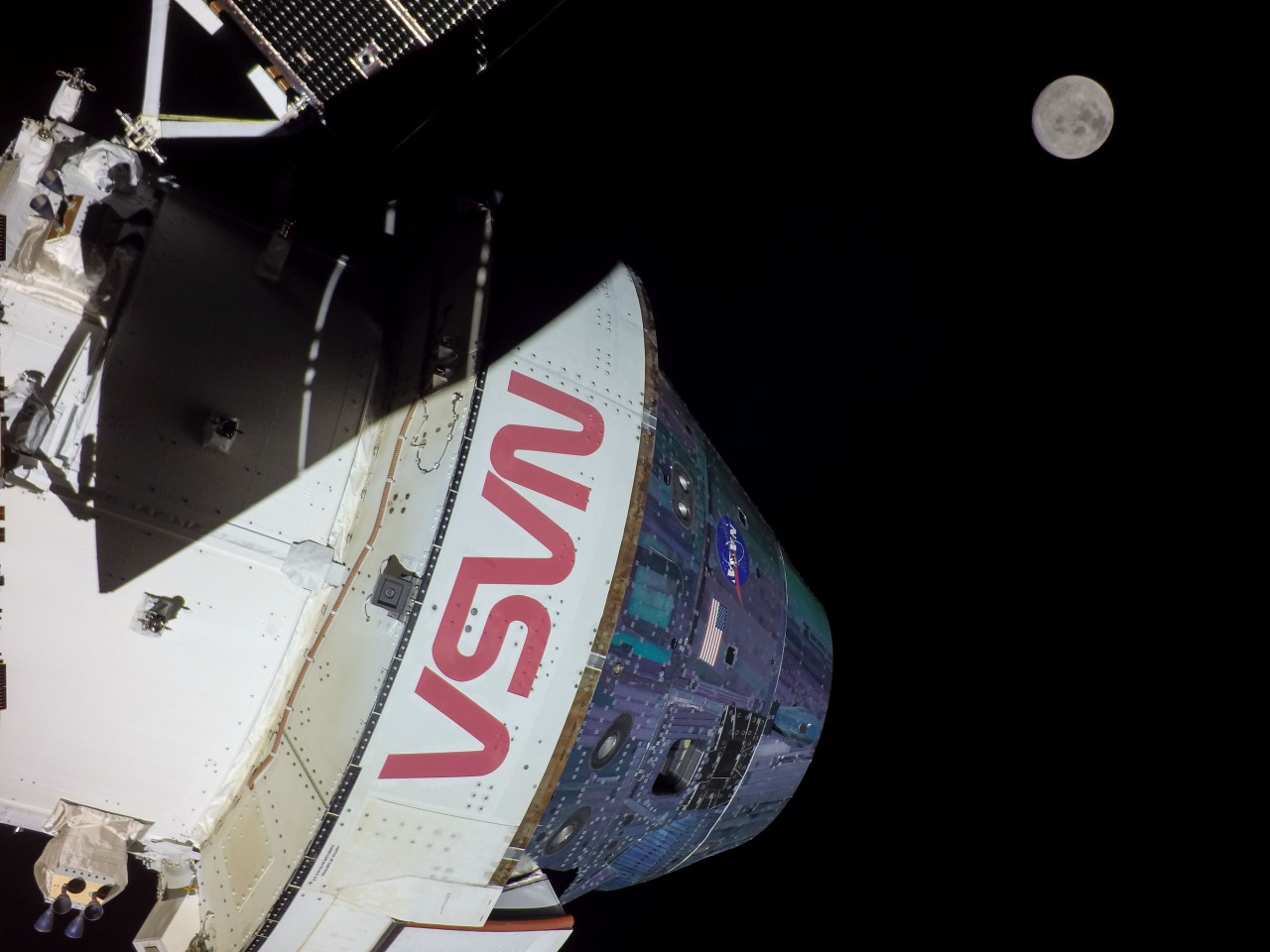 NASA’s Orion spacecraft in space looking at the Moon from a camera mounted on one of its solar arrays. The Orion spacecraft appears in the foreground. The Earth and the Moon appear in the far distance against the blackness of space. The Moon appears just slightly larger than Earth. Credit: NASA