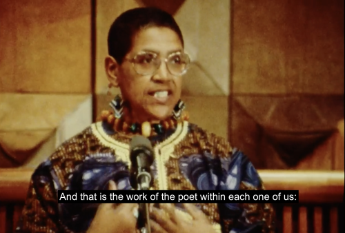 Audre Lorde stands at a podium accepting her award and appointment as State Poet of New York for 1991-1993. She is dressed formally in a colorful tunic. She raises both hands to her chest and continues, "And that is the work of the poet within each one of us:"