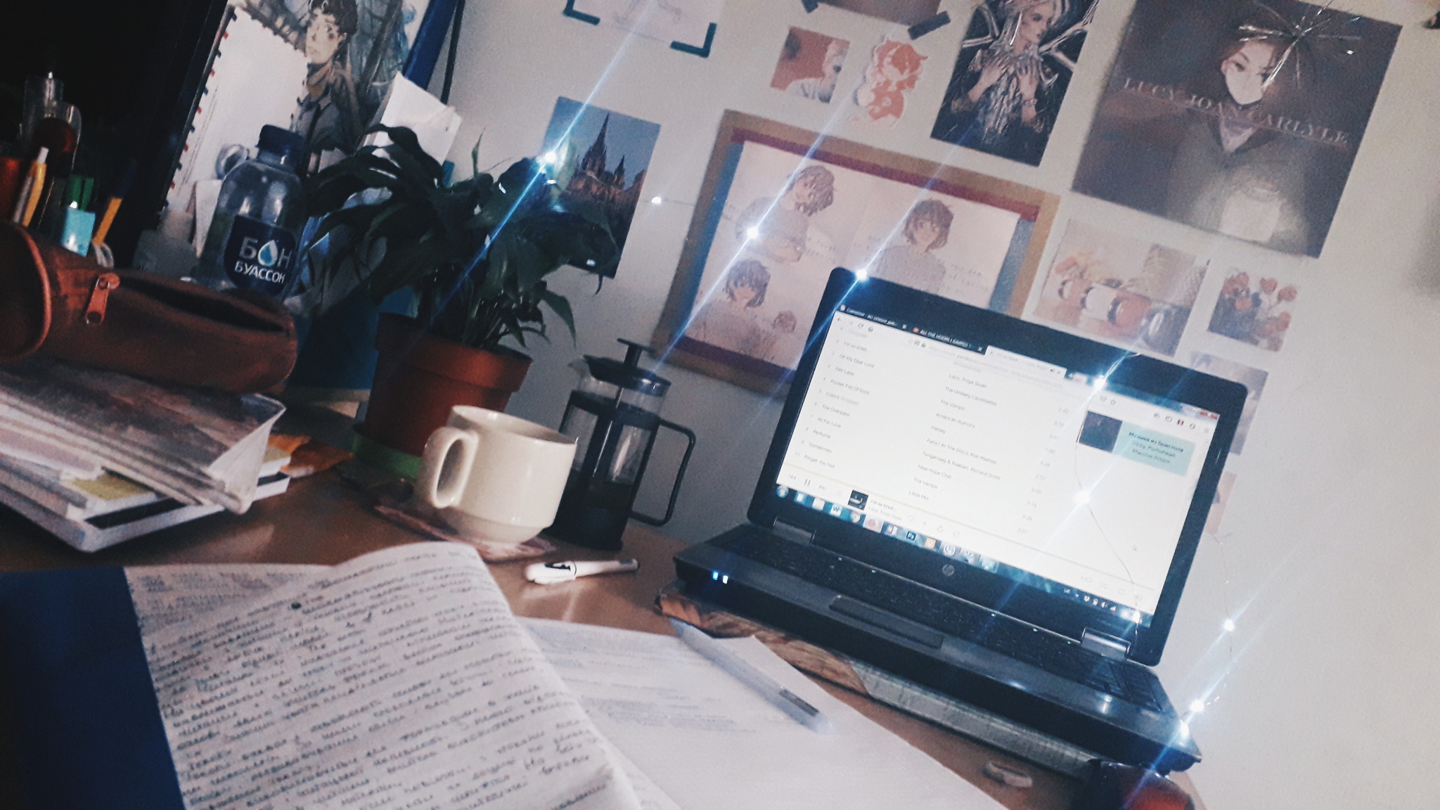 a photo of a desk with a laptop on it, a plant, fairy lights, notebooks, a French press and a cup. On a blue wall there are a lot of small pictures 