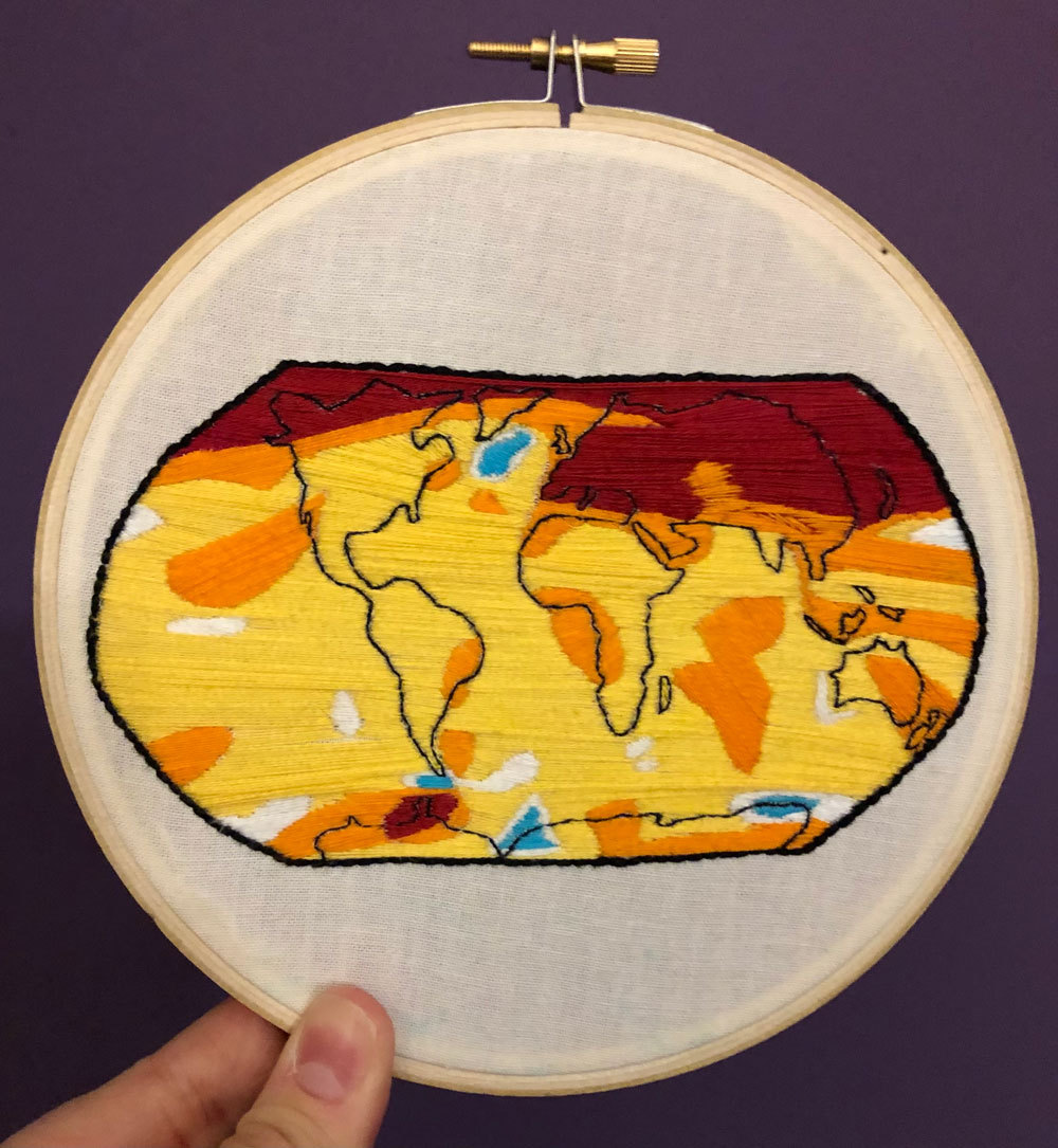 This image shows an embroidered art piece based on NASA’s yearly temperature release. To the bottom left, two fingers hold up the circular piece. A round wooden frame holds it in place. In the center, a map appears of the different content. It’s outlined in black. Most of the map is covered in yellow stitching to show a warming pattern. To the left and right, the stitches change to an orange color and are scattered on the map. In the top left- and right-hand corners, the color changes to a dark red to signify another temperature change.