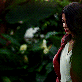 Isabelle (Emeraude Toubia) wearing a red leather jacket over a white top, turns around in front of greenery