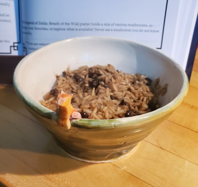 a bowl of mushroom risotto. The bowl is moss green with a white interior, on the lip sits an orange fox adjacent ceramic figure.