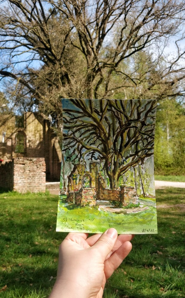 A photo taken outside, showing a hand holding up a small watercolour painting. The painting is of a large tree with many branches but without any leaves. It stands tall next to the ruin of a chapel, with only a few brick walls remaining. A couple of pieces of cloth are tied to some of the lower branches. Behind the painting the same chapel and tree is visible, surrounded by greenery and bathed in bright sunlight.