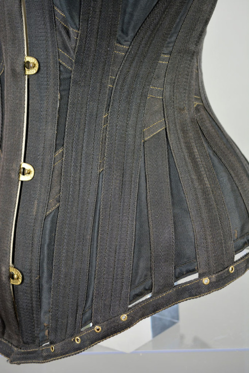 This black lasting corset is lined with white cotton twill and edged with gold and black braid. It w