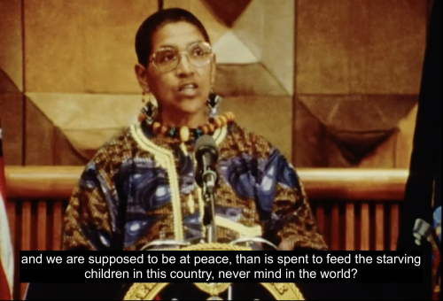 Audre Lorde stands at a podium accepting her award and appointment as State Poet of New York for 1991-1993. She is dressed formally in a colorful tunic. She continues, "I've been asking myself, 'and we are supposed to be at peace, than is spent to feed the starving children in this country, never mind in the world?'"