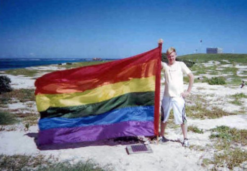 A man standing in the dunes of an island proudly holding up a rainbow flag as tall as him attached to a pole.