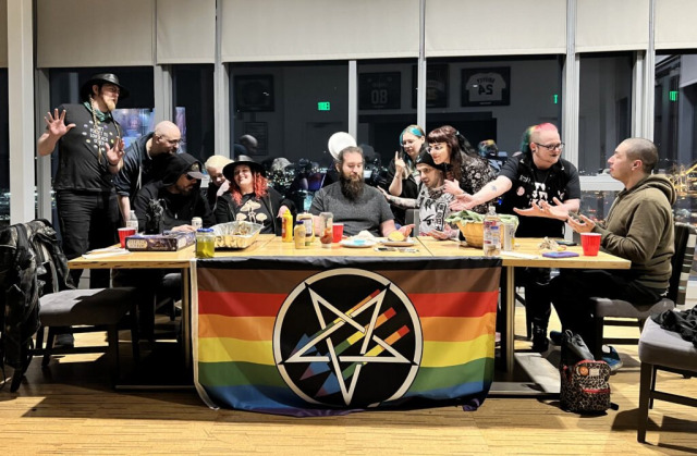 a menagerie of satanists reproduce "the last supper" at a modern table. On the front of the table is a gay pride antifascist pentagram flag.