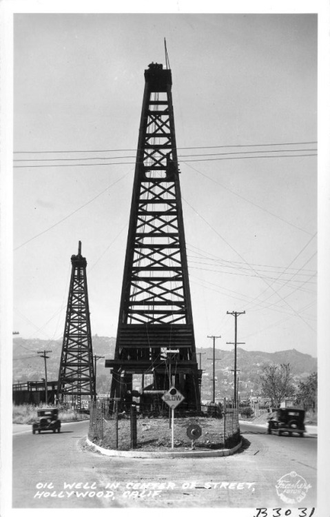 Cars pass a towering oil well in the center of a Hollywood street (possibly La Brea Avenue), 1936.