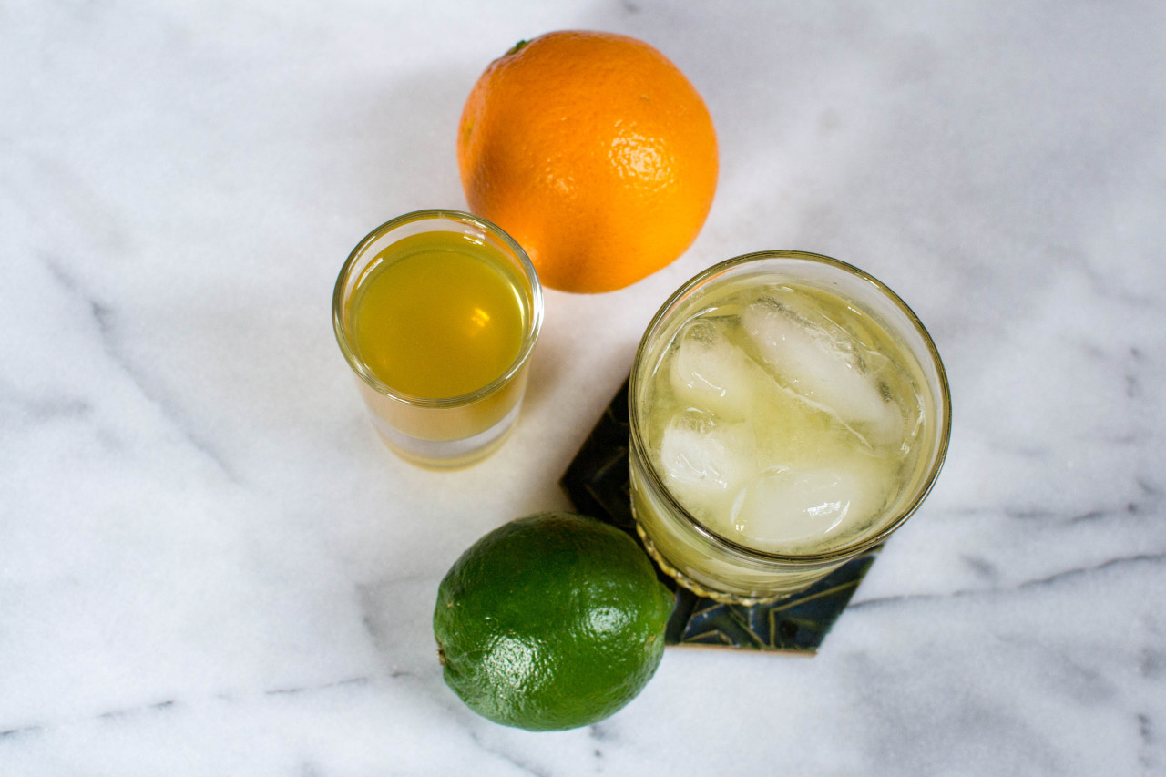 Overhead photograph of a margarita in a crystal glass on a hexagonal coaster, with a lime next to it and a shot glass of orange liqueur next to an orange behind them, all on a marble background