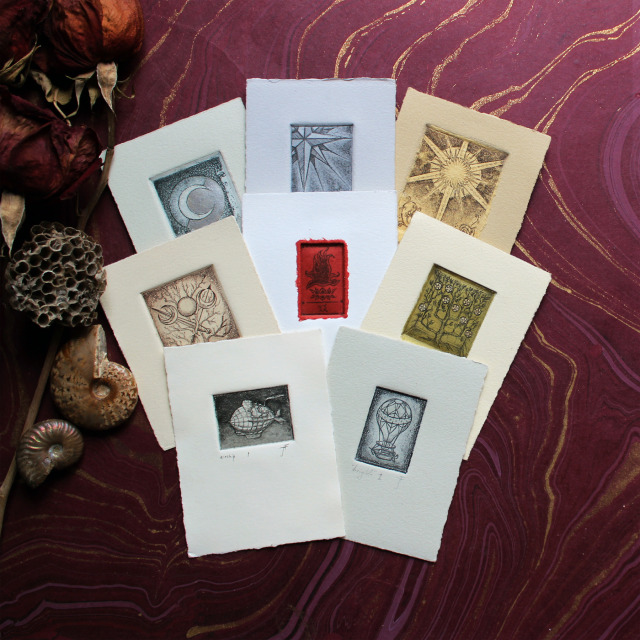 A photo of eight 3x4” hand-embellished etchings fanned out on a wine colored sheet of marbled paper. The imagery of the etchings resembles tiny, enigmatic tarot cards.