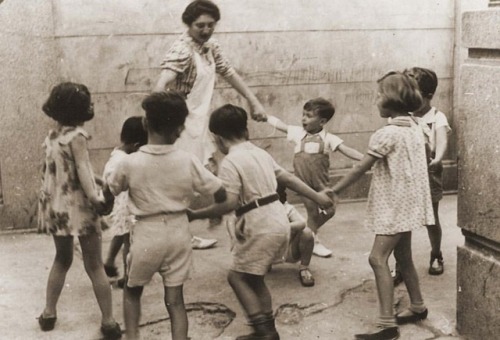 historicity-was-already-taken: A teacher leads a group of young children in a circle dance at the Ka