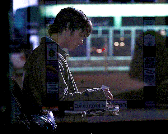 a gif of sam winchester, from season one of supernatural. they're seen sitting in a motel lobby, through the glass of the window. they read something held in their lap, briefly, then turn their head and face towards the camera.
