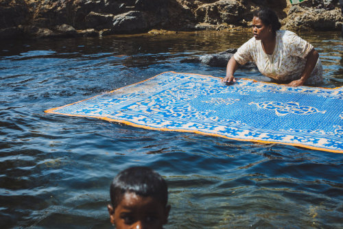 brassmanticore:A Nubian woman cleaning her carpet in the Nile while her child is having a swim.Nour 