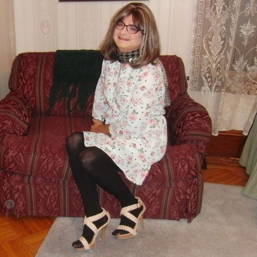 Full-length picture of me, seated, knees right, wearing my new purple cat-eye glasses, floral shirt 
