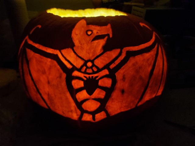 Front view and whole view of a pumpkin carved so that its flesh glows bright orange without being all the way cut through. The image is of a Mesoamerican styled image of a bat, wings outspread, with an open rib cage showing the heart. The circular eye is slightly brighter than the rest of its body.