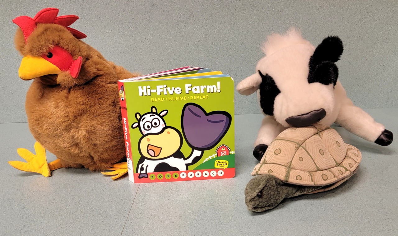 A stuffed chicken, turtle, and cow gathered around the book which shows a cow raising a hoof to give a high five