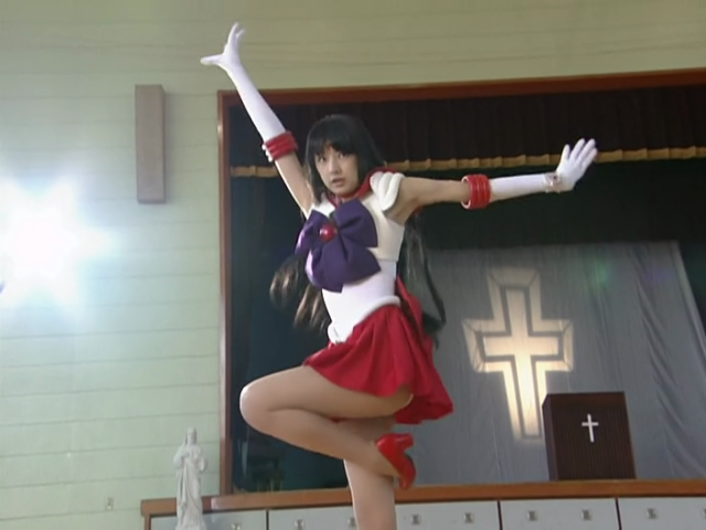 Sailor Mars (played by Keiko Kitagawa) striking a somewhat ridiculous pose with arms raised and knee bent, kinda like a bird. Behind her is a stage in some kind of large gym auditorium, upon which stands a pulpit adorned with a white cross, and behind which is projected the image of another large spinning cross.