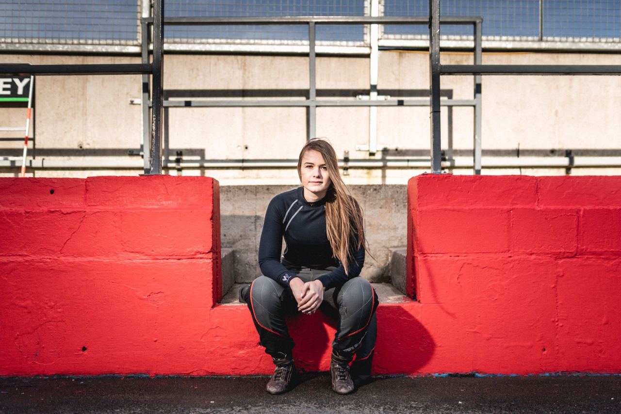 Cristiana Oprea, a racer, wears a black Walero racing undergarment while sitting on a red divider at the edge of a racetrack.