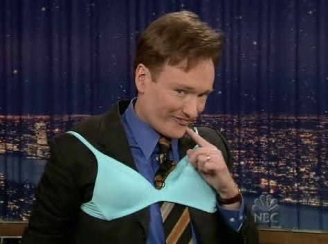 Conan O'Brien wearing a teal bra over his suit and tie. He is pointing a finger to the corner of his mouth with a silly grin.