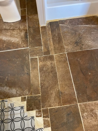 a bathroom floor. large brown tiles have been haphazardly cut into strips and little squares to fit. mismatched tiny white tiles encroaches in a semicircle from the corner.