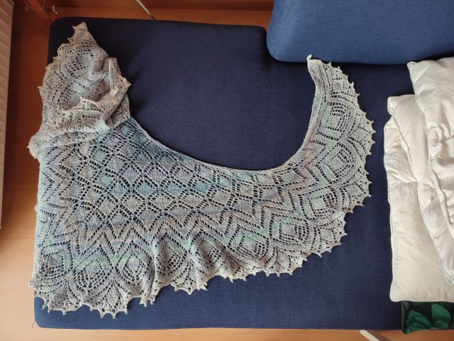 A light blue handknit lace shawl, draped over a dark blue sofa for display. It's slightly crumpled and distorted.