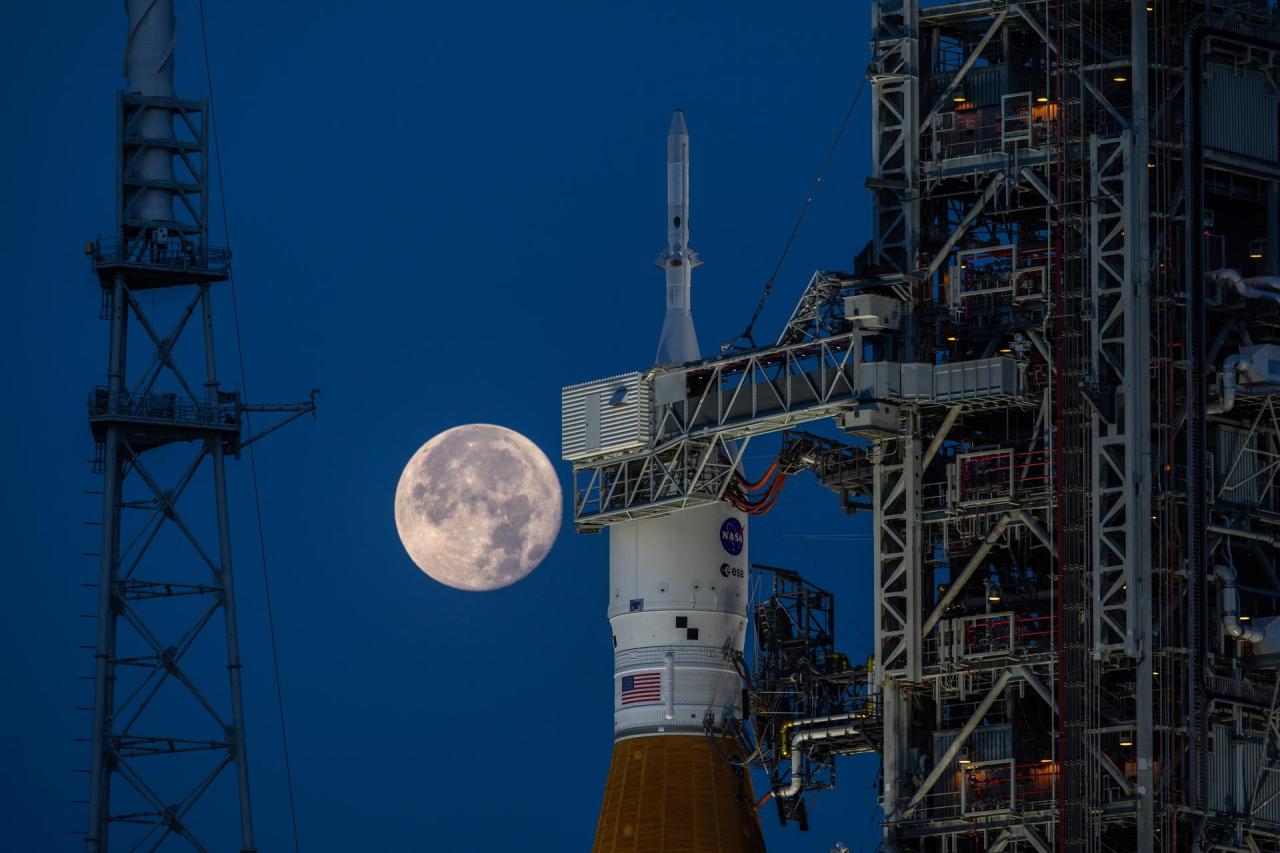 The SLS and Orion spacecraft can be seen in the foreground with a full Moon to the left of the spacecraft. The SLS is orange, Orion is white, and the Moon is grey and white. A lightning tower is to the left of the Moon.  Credit: NASA/Ben Smegelsky