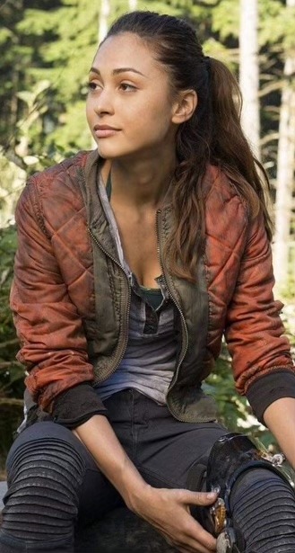 An image of Raven Reyes from the live action TV show The 100. She is in the frame from the knees up, sitting on a log. She is looking to the left of the frame with a slight smile, she has her hands on a brace on her right knee. She is wearing a gray shirt with a dirty orange semi puffy jacket over it. The jacket has black cuffs and is green around the zipper, it is unzipped. She is wearing black jeans. She has elbow length brown hair, some of it is up in a ponytail. She has a medium light skin tone. The background is blurred, but of a forest.