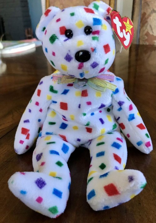a white bear plush scattered with multicolored confetti patterns and donning a pink, yellow, and blue ribbon bowtie.