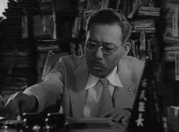 A scene from Ikiru (1952) by Akira Kurosawa. A man shuffles papers on his desk with the caption, "Busy. Always so very busy. But in fact, this man does nothing at all."